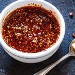 A bowl of chili crisp sauce placed next to a spoon with a small piece of butter, captured on a dark rustic background.